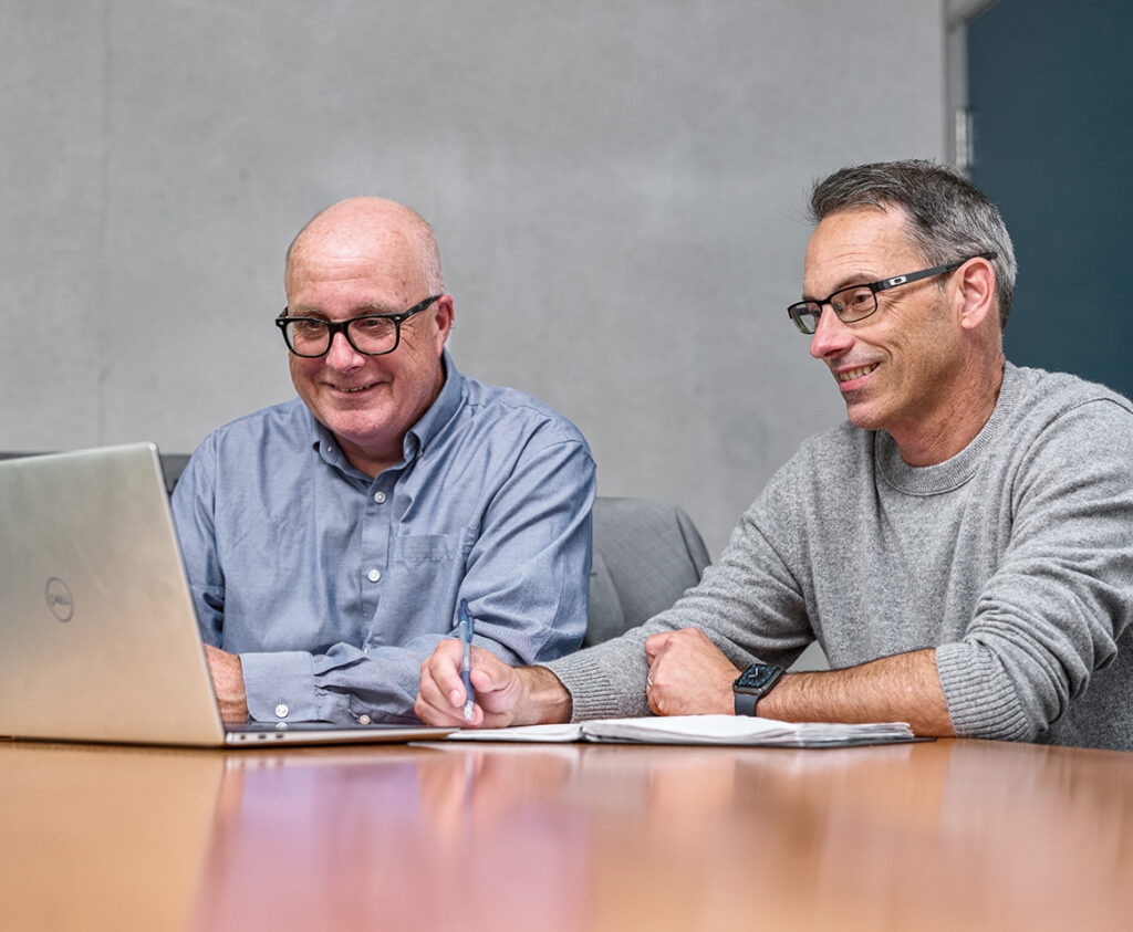 Two businessmen looking at a laptop and taking notes with a pen.